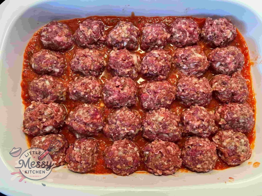 Baking dish with marinara sauce topped with ground meat meatballs, ready for baking.