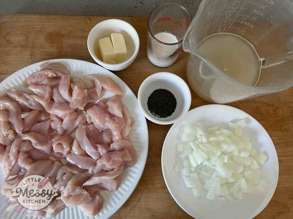 Ingredients set on a cutting board, including, sliced chicken breast, diced onions, dill, butter, cream and chicken broth.