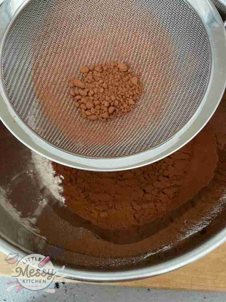 Sifting cocoa powder through a mesh strainer into a mixing bowl to remove lumps.