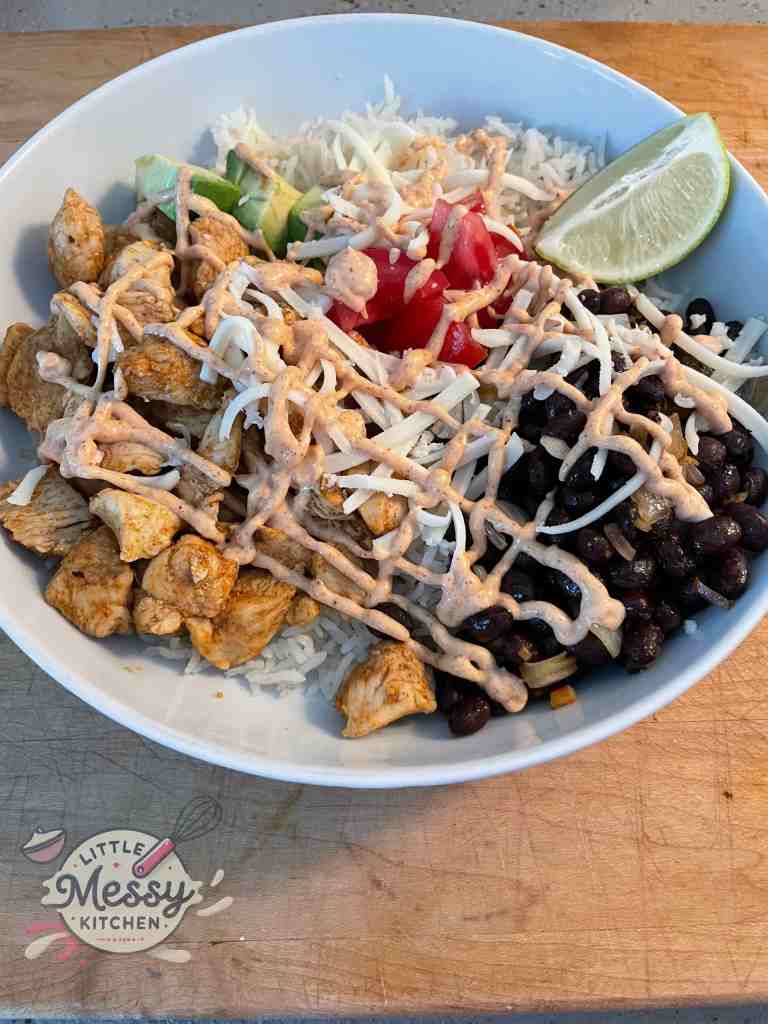 Marinated chicken, black beans, avocado, tomato and slice of lime on a bed of rice with taco sauce drizzled on top.