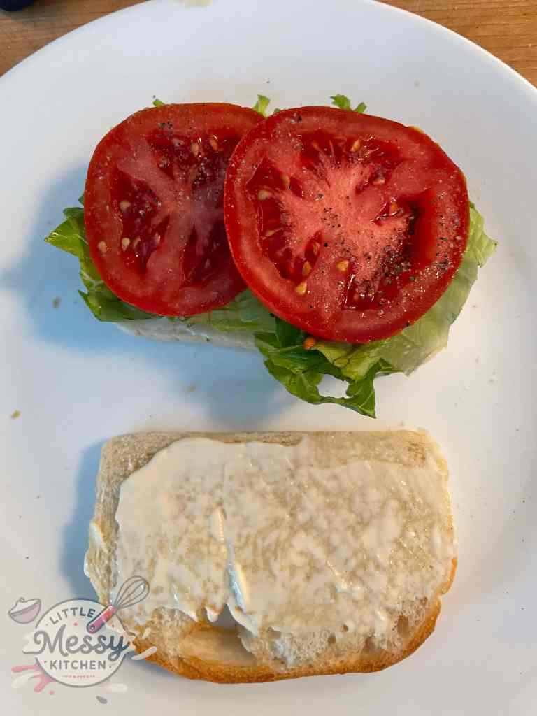 Open-faced sandwich with one slice of bread topped with lettuce and two slices of seasoned tomato, and the other slice spread with mayonnaise, ready for assembly.