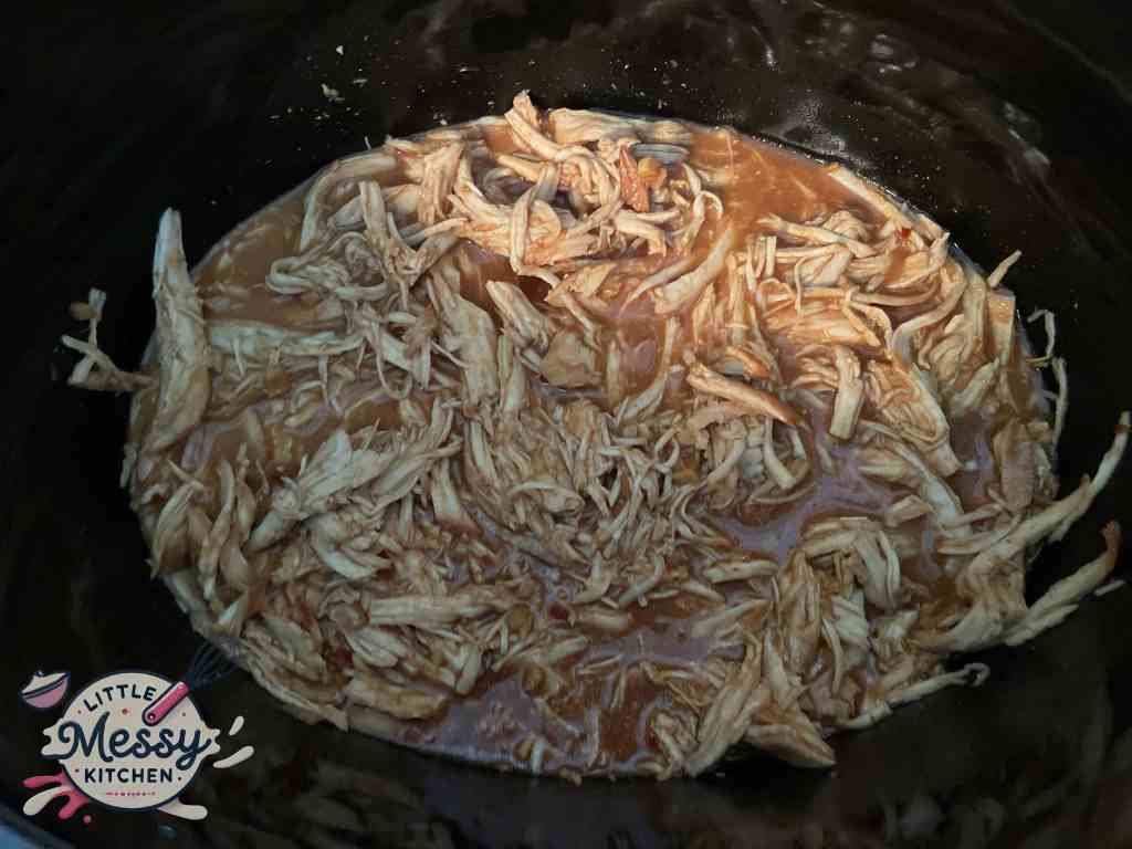 Shredded cooked chicken breast mixed with sauce in a slow cooker.