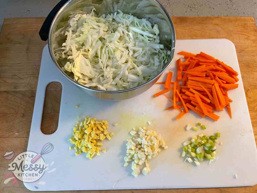 cabbage shredded in a bowl along side minced garlic, ginger and green onions and matchsticks carrots on a cutting board