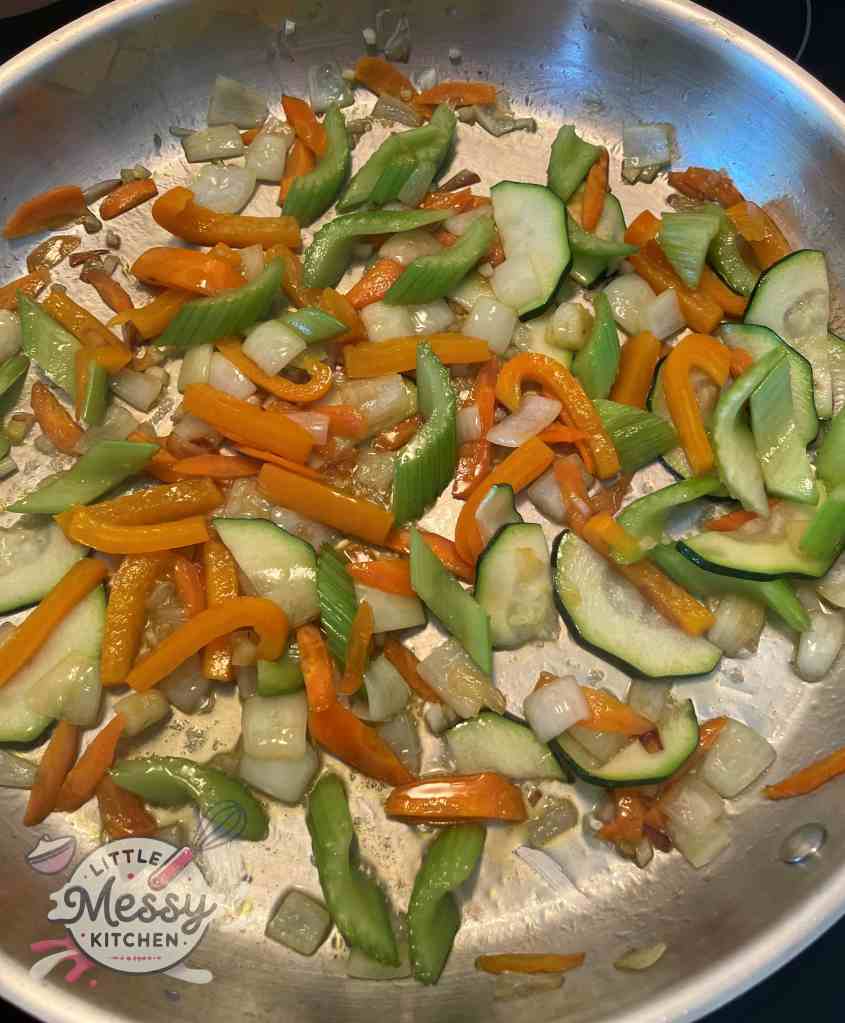 Chopped vegetables including sliced carrots, celery, bell pepper, zucchini, diced onion, and minced garlic, sauteing in frying pan