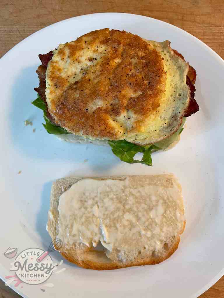 Open-faced sandwich with one slice of bread topped with lettuce, tomato, bacon, and a round fried egg, and the other slice spread with mayonnaise, ready for assembly.