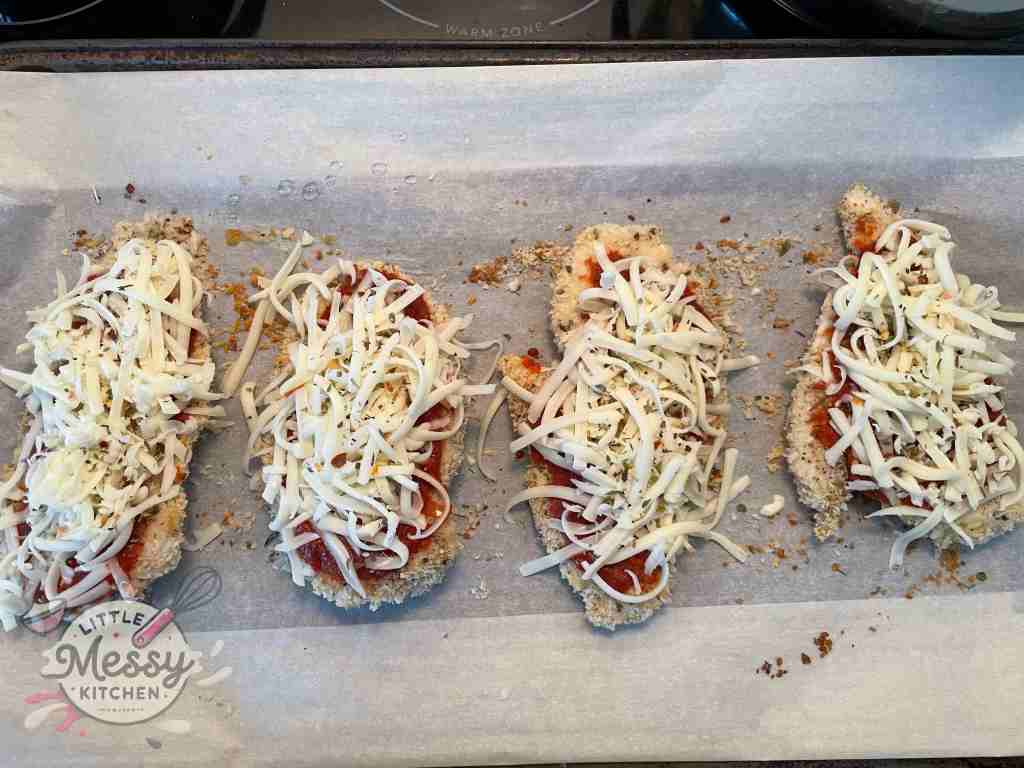 Breaded chicken breast topped with marinara sauce and cheese on a baking sheet ready to be cooked.