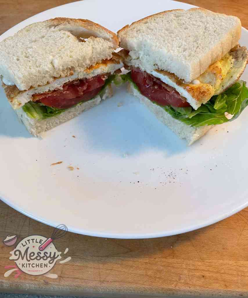 Completed BELT sandwich cut in half, showing layers of lettuce, tomato, bacon, and egg between slices of bread, served on a white plate.