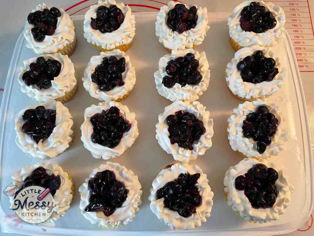 16 cupcakes topped with whipped cream and blueberry mixture arranged on a tray.
