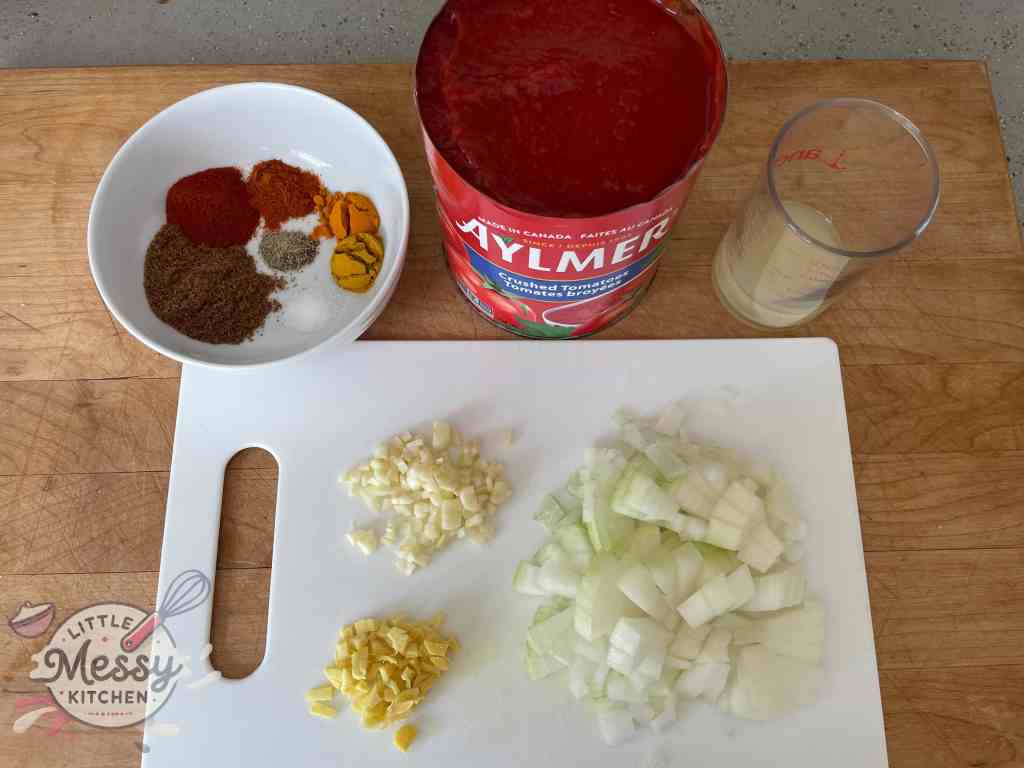 Ingredients set on a cutting board, including: crushed tomatoes, Lemon juice, spices, onions, garlic and ginger.
