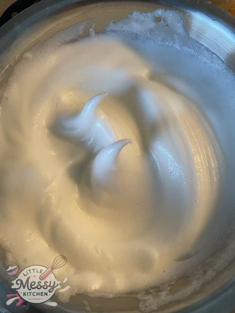 Egg whites whisk to stiff peaks in a mixing bowl.